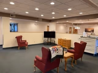 A cozy common area with four red upholstered armchairs arranged around a wooden coffee table. A flat-screen TV is mounted on a stand against a half wall, with a wooden dresser nearby. The room has a carpeted floor, recessed ceiling lights, and light-colored walls with minimal decorations.