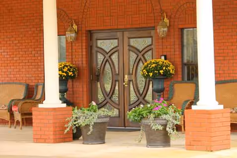 Brick building entrance with double wooden doors, white columns, chairs, and potted flowers on the porch.