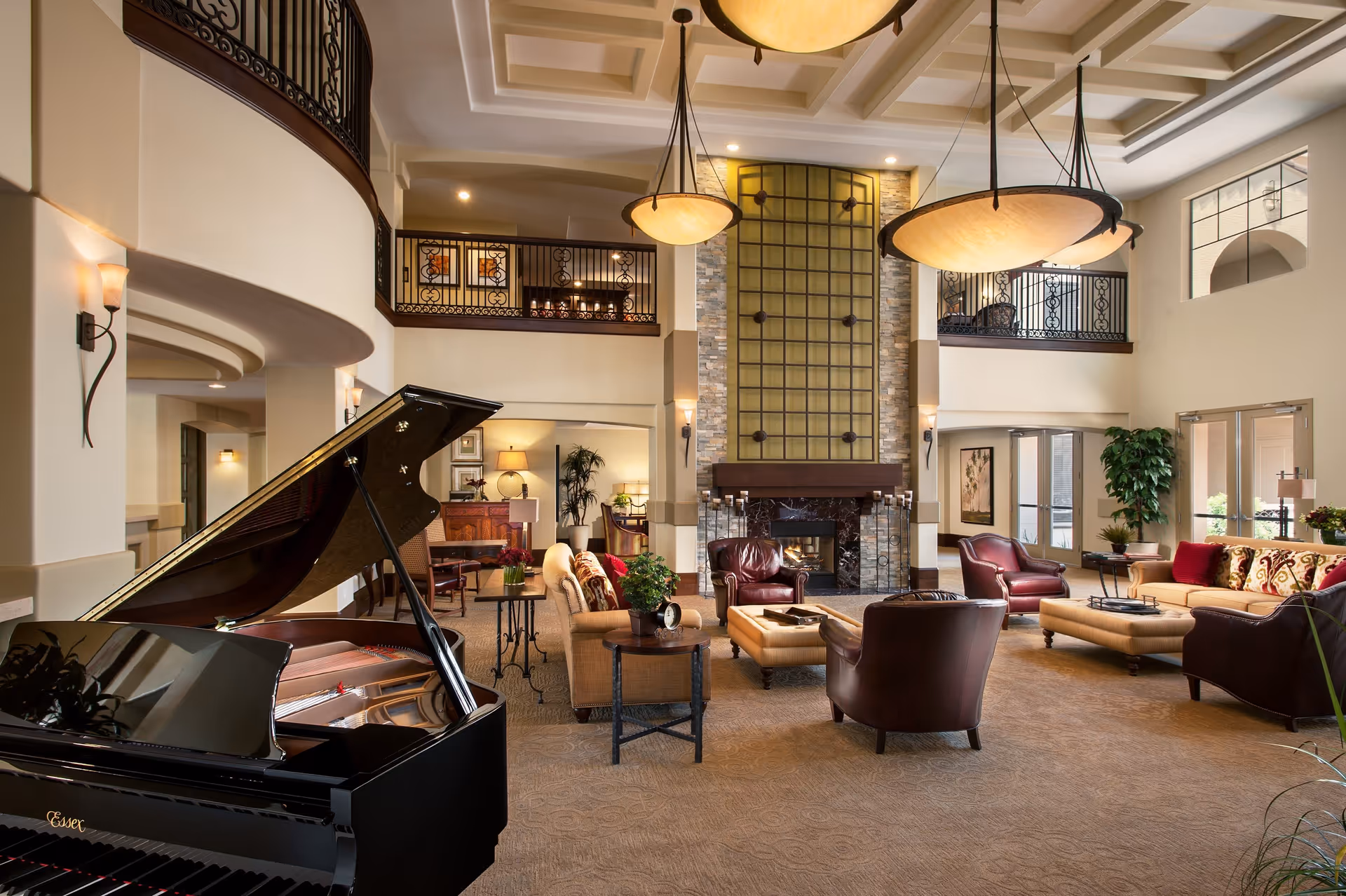 Spacious and elegant living room with high ceilings, large hanging light fixtures, a grand piano, comfortable seating including sofas and armchairs, a stone fireplace with decorative metal grid above it, and large windows letting in natural light.