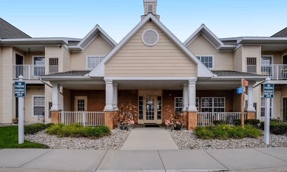 Front exterior view of Devonshire Retirement Village building with a covered entrance supported by white columns, beige siding, brick accents, and landscaped areas with plants and rocks on either side of the walkway.
