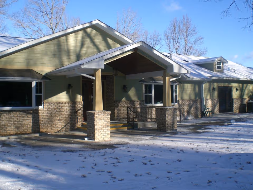 Exterior view of a single-story building with green siding and brick accents, featuring a covered entrance supported by two brick pillars. The ground is covered with a light layer of snow and leaf debris, and leafless trees are visible in the background under a clear blue sky.