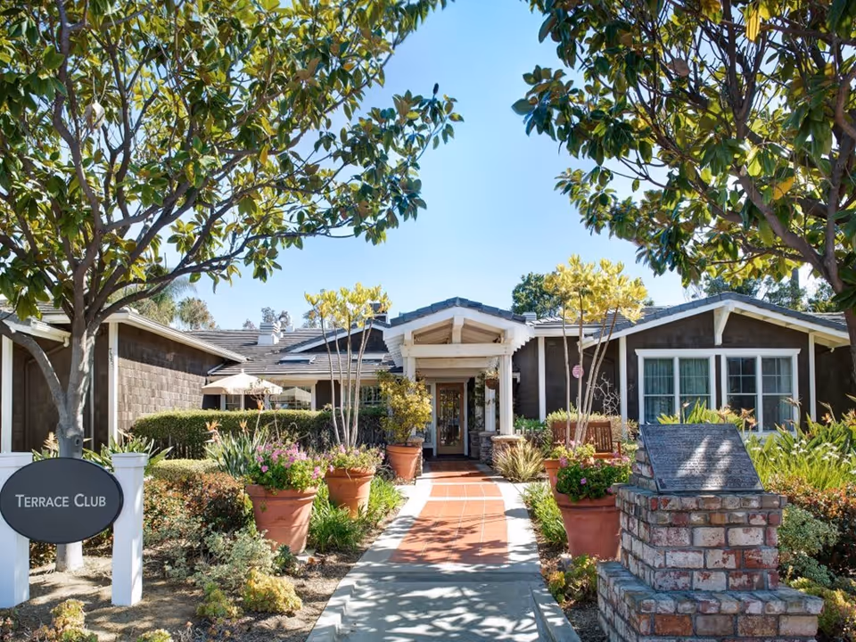 Exterior view of a single-story building with a tiled walkway leading to the entrance, surrounded by potted plants, trees, and well-maintained landscaping under a clear blue sky. A sign near the walkway reads 'Terrace Club.'