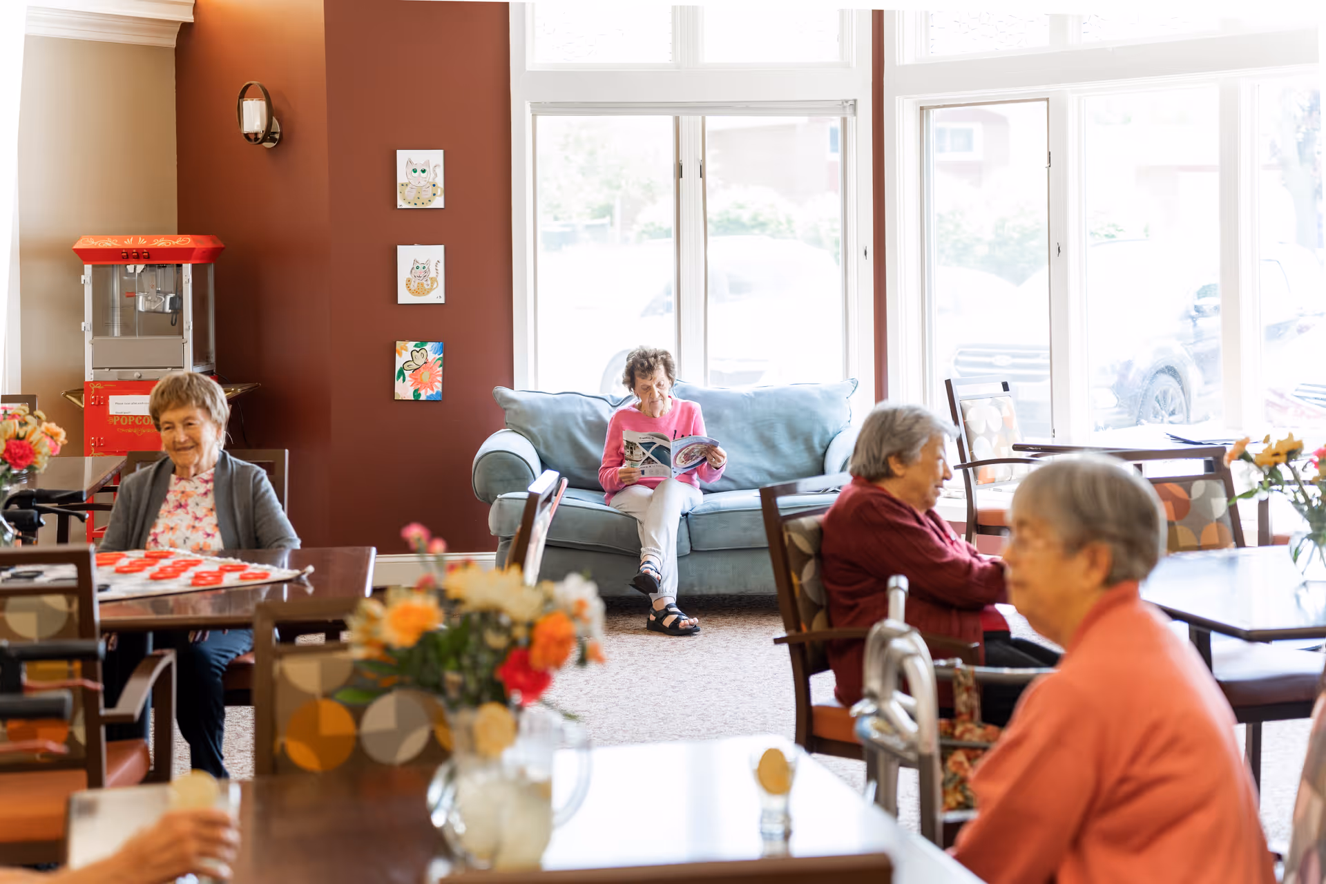 A bright common area in a senior living community with several elderly women sitting at tables and a woman sitting on a light blue couch reading a magazine. The room has large windows letting in natural light, a popcorn machine in the corner, and colorful artwork on the wall.