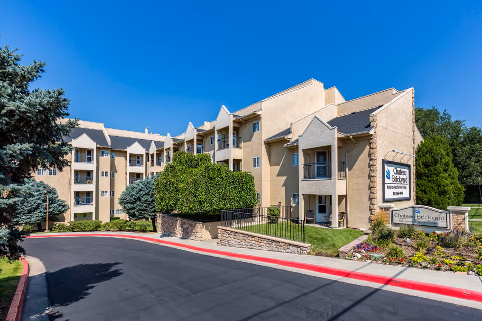 Three-story senior living building with balconies, landscaped entrance, and a sign reading "Chateau Brickyard" under a clear blue sky.