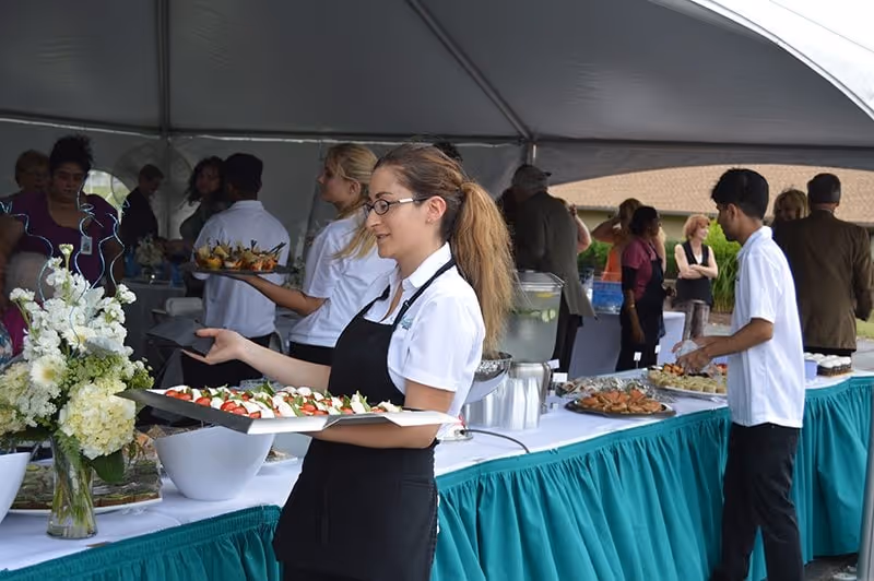 A catering event under a large white tent with several servers dressed in white shirts and black aprons serving trays of appetizers and food to guests. There is a long table covered with a white tablecloth and teal skirt, decorated with a large floral arrangement and various platters of food and beverages. People are gathered around the table, selecting food and socializing.