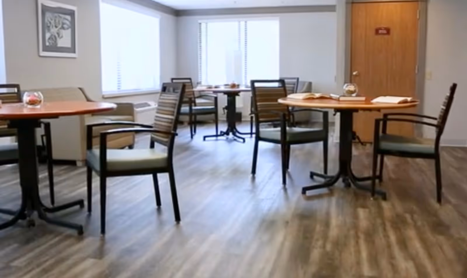 A bright room with several round wooden tables and chairs arranged around them. There are windows with blinds letting in natural light, a beige couch against the wall, and a wooden door in the background. The floor has a wood-like finish and the walls are painted white.