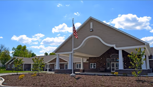 Front exterior view of a senior living facility building with a covered entrance, an American flag on a flagpole, small trees, and a clear blue sky with some clouds.