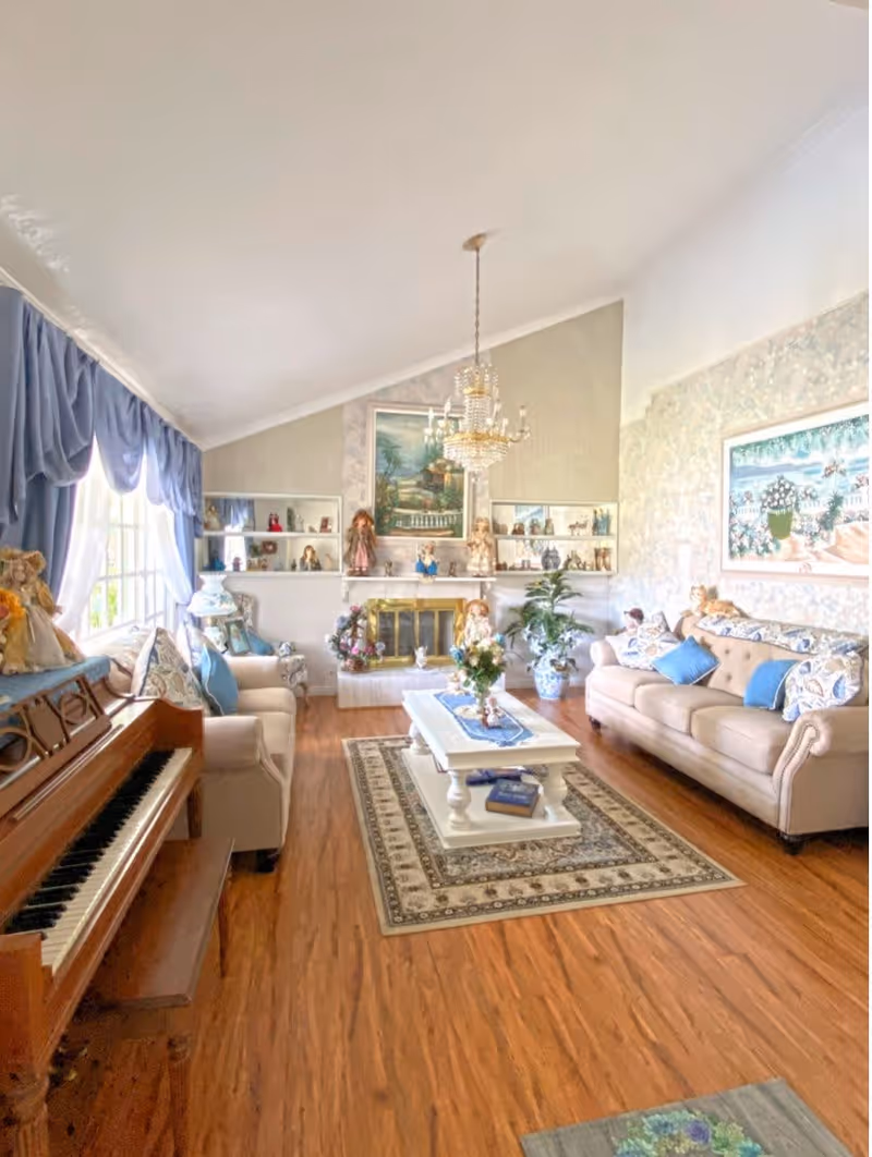 A bright and cozy living room with wooden flooring, two beige sofas adorned with blue and patterned cushions, a white coffee table with decorative items, a piano on the left side, and a fireplace with shelves displaying various dolls and figurines. The room has large windows with blue curtains, a chandelier hanging from the ceiling, and framed artwork on the walls.