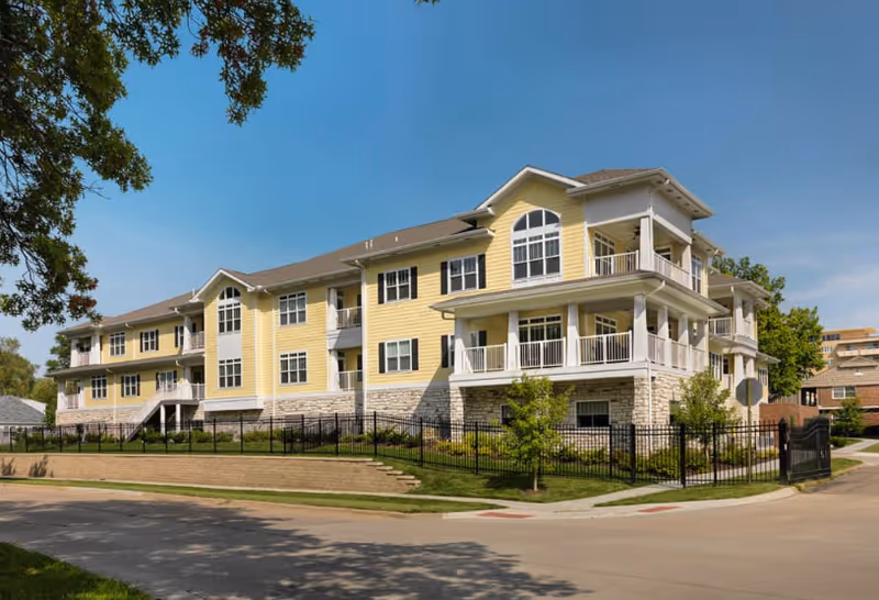 Exterior view of a three-story senior living facility building named Brewster Place, painted yellow with white trim, featuring multiple balconies and large windows, surrounded by a black metal fence and greenery under a clear blue sky.