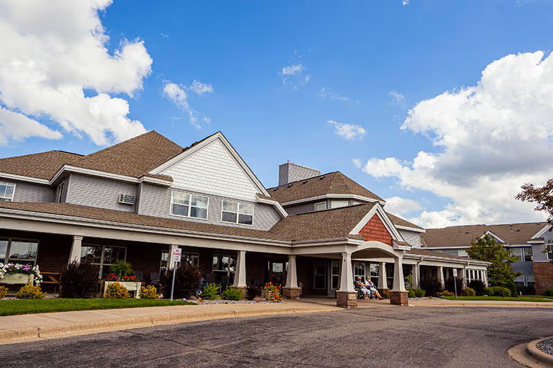 Exterior view of Guardian Angels By The Lake Senior Living facility in Elk River, showing a large building with a covered entrance, multiple windows, and a driveway under a partly cloudy sky.