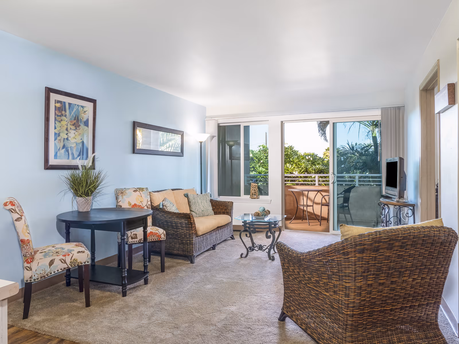 A bright living room with light blue walls, featuring a wicker sofa with beige cushions, a wicker armchair, a small glass coffee table with decorative items, and a black round table with two floral upholstered chairs. There are two framed artworks on the wall and a floor lamp. Large sliding glass doors open to a balcony with outdoor seating and lush greenery visible outside.