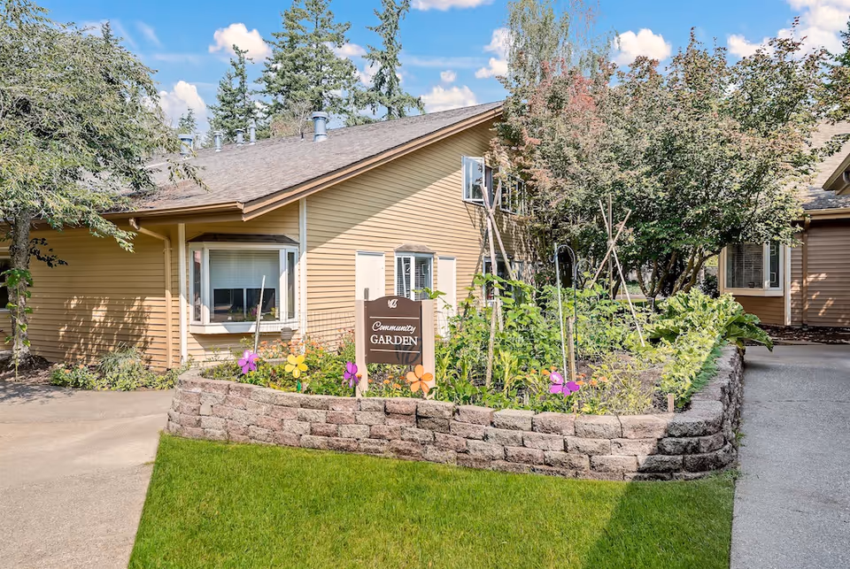 Exterior view of a single-story beige building with a raised community garden bed and a sign reading 'Community Garden'.