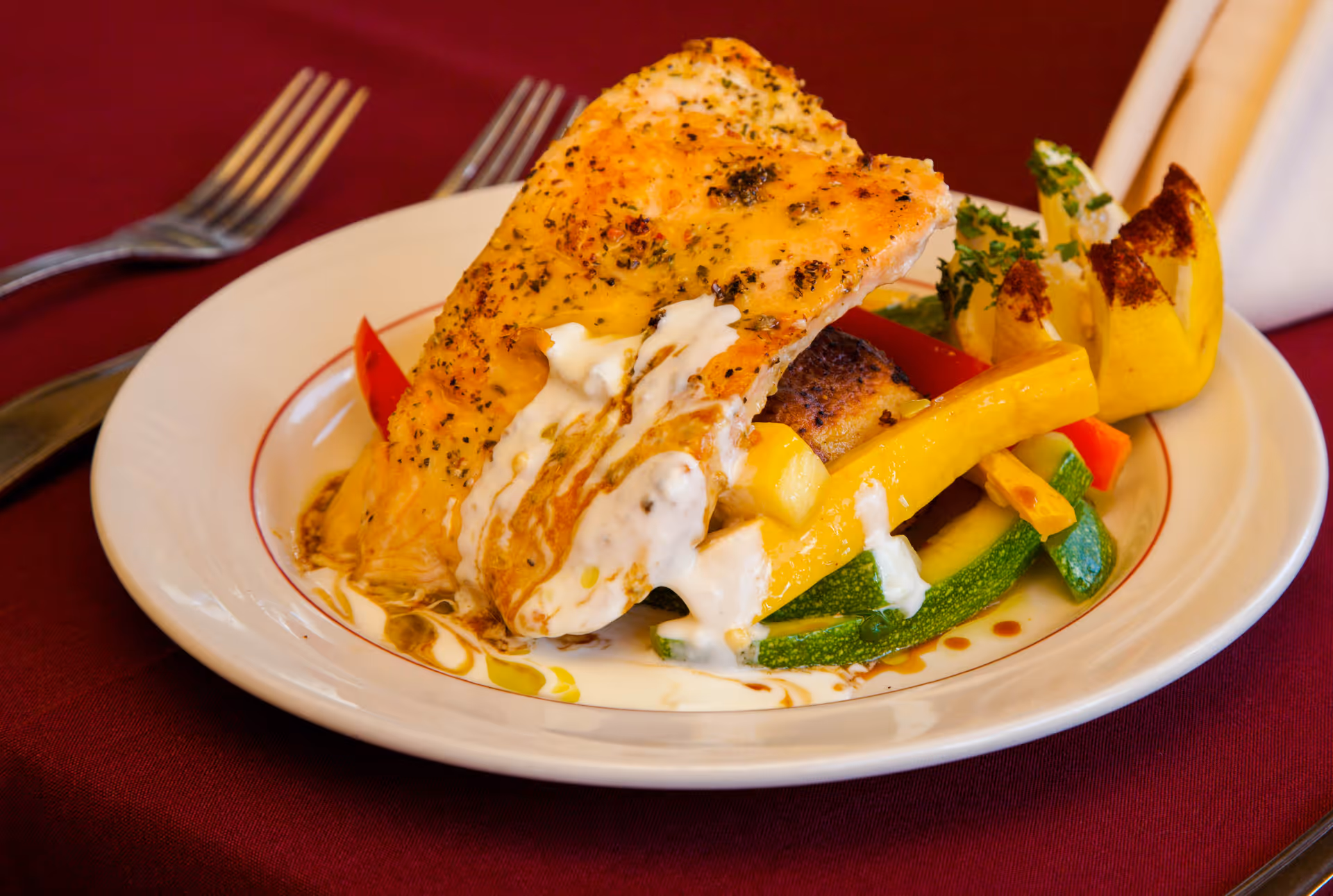A plated dish featuring a seasoned and cooked piece of fish topped with a creamy white sauce, served on a bed of mixed sautéed vegetables including yellow squash, zucchini, and red bell peppers, with a decorative lemon garnish on the side. The plate is set on a burgundy tablecloth with a fork and knife placed nearby.