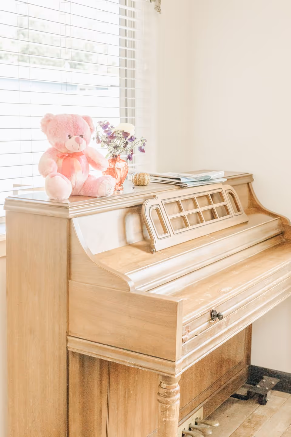 A light wooden upright piano placed against a wall near a window with white blinds. On top of the piano, there is a pink teddy bear with a pink ribbon, a small orange vase with purple and white flowers, a small decorative pumpkin, and some folded newspapers.