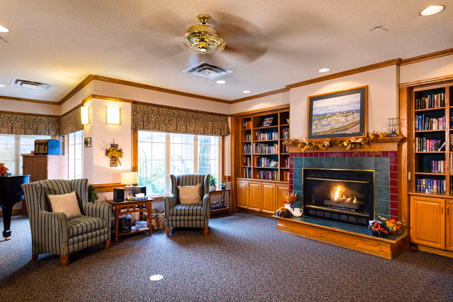 A cozy living room in an assisted living facility featuring two striped armchairs with cushions, a wooden side table with a lamp and stereo equipment, a lit fireplace with a decorative mantle, built-in wooden bookshelves filled with books, a large window with patterned valances, and a ceiling fan overhead.
