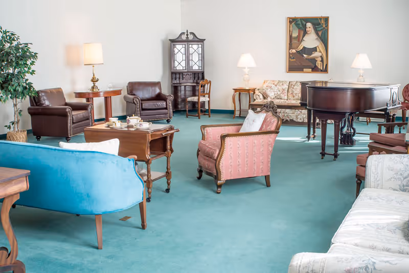 A spacious living room with vintage-style furniture including a blue sofa, pink armchair, brown leather chairs, a floral couch, wooden tables with lamps, a grand piano, and a framed portrait on the wall. The room has a teal carpet and white walls.