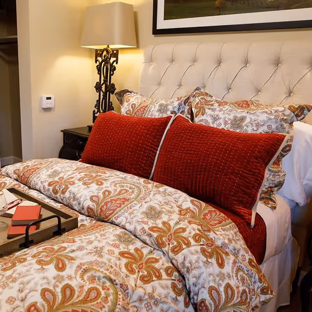 A cozy bedroom featuring a bed with a tufted beige headboard, decorated with patterned pillows and a comforter in shades of red, orange, and cream. Two large red textured pillows are placed in front of the patterned pillows. A wooden tray with a book and glasses rests on the bed. A decorative lamp with a beige shade sits on a dark nightstand beside the bed, and a thermostat is mounted on the wall.
