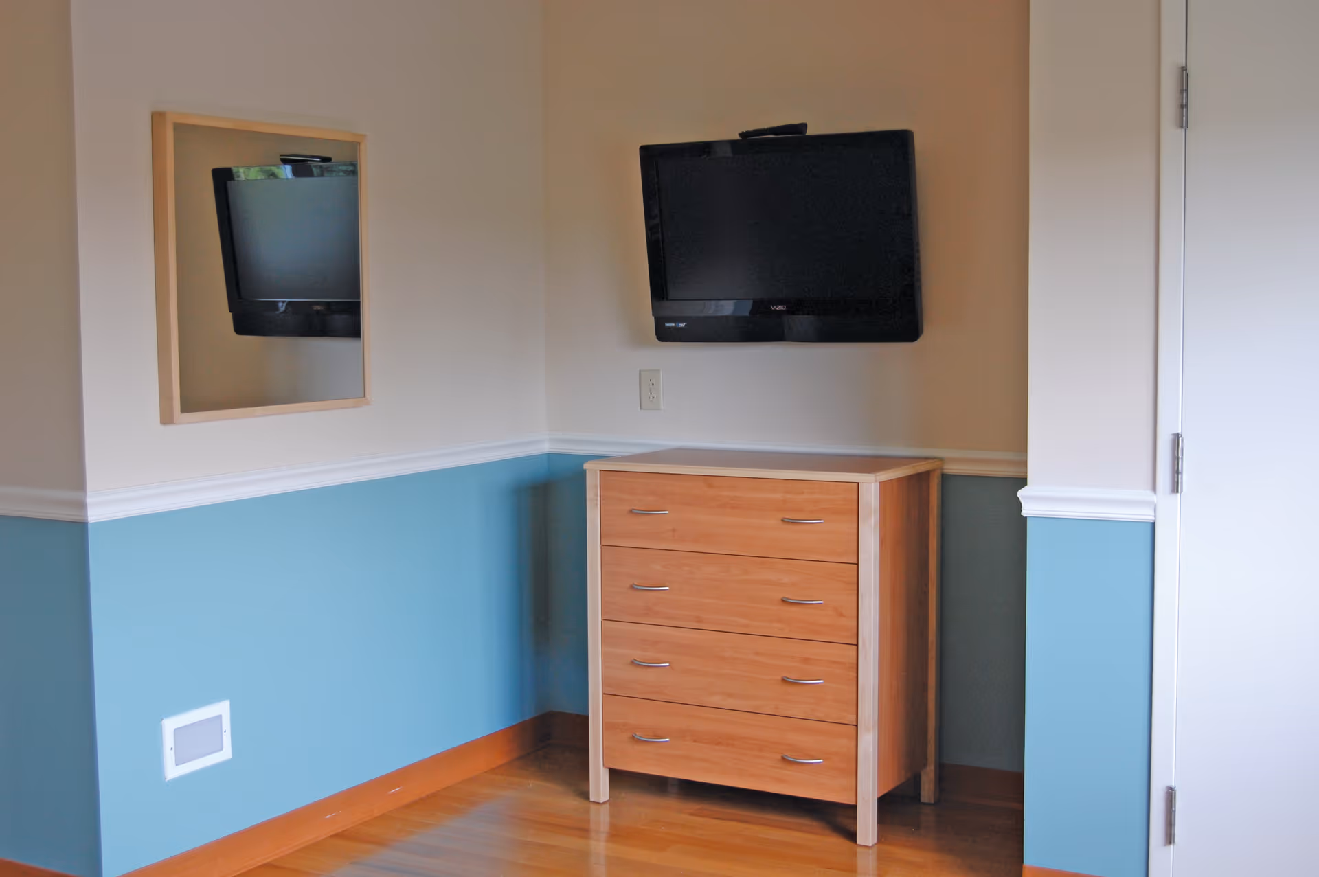 A corner of a room with light blue and beige walls, featuring a wooden dresser with four drawers, a flat-screen TV mounted on the wall above the dresser, a mirror on the adjacent wall reflecting the TV, and a white door on the right side.