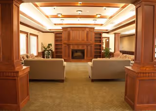 A spacious living room with beige sofas arranged facing a wooden fireplace with built-in shelves. The room features wooden columns, recessed ceiling lights, large windows with blinds, and green carpeted flooring. There are plants placed near the windows and in the corners of the room.