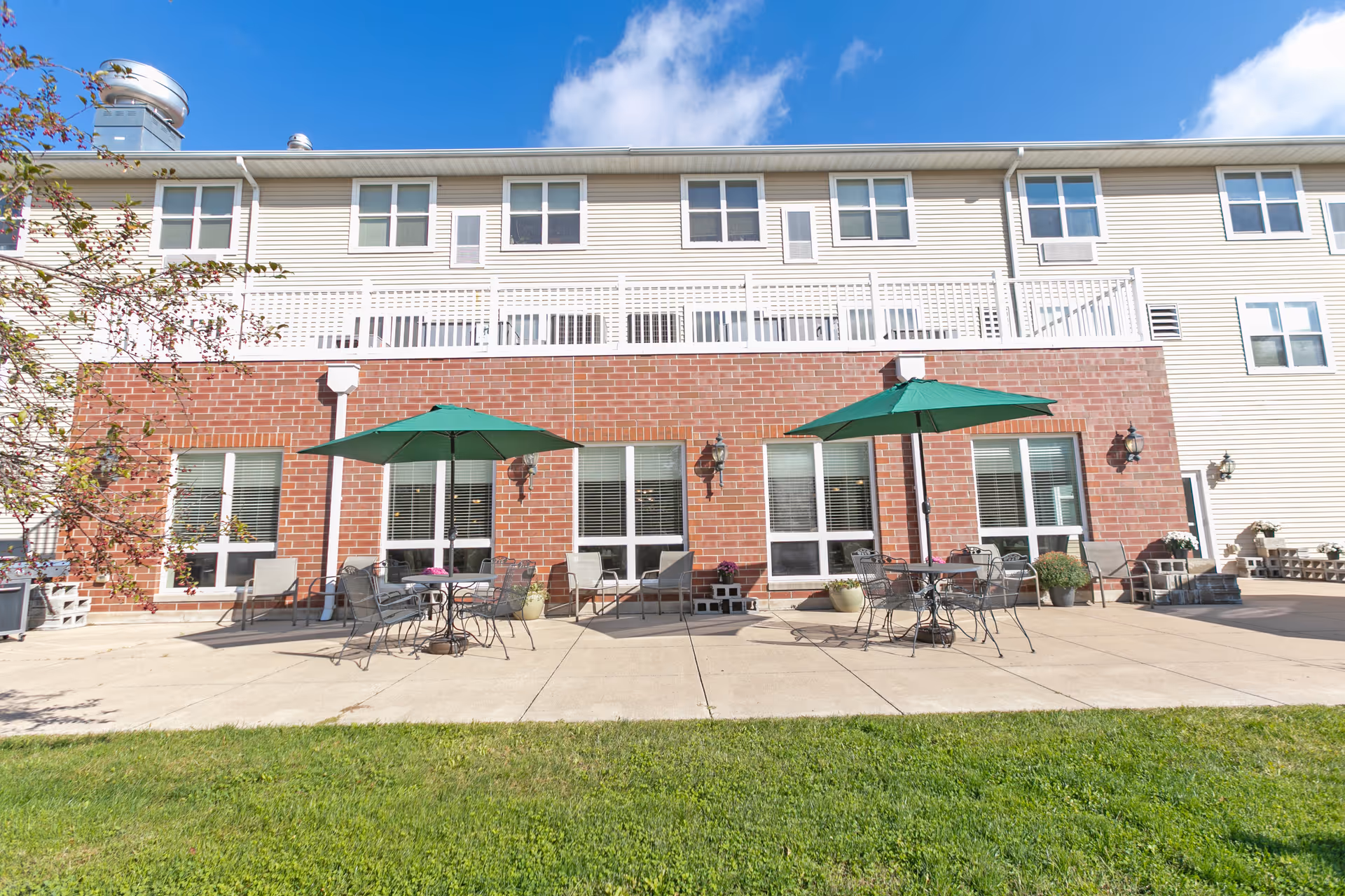 Outdoor patio area of a senior living facility with two green umbrellas shading metal tables and chairs. The building has a brick lower facade with large windows and a beige upper facade with multiple windows. There are potted plants and outdoor lighting fixtures on the wall, with a grassy lawn in the foreground under a blue sky with some clouds.