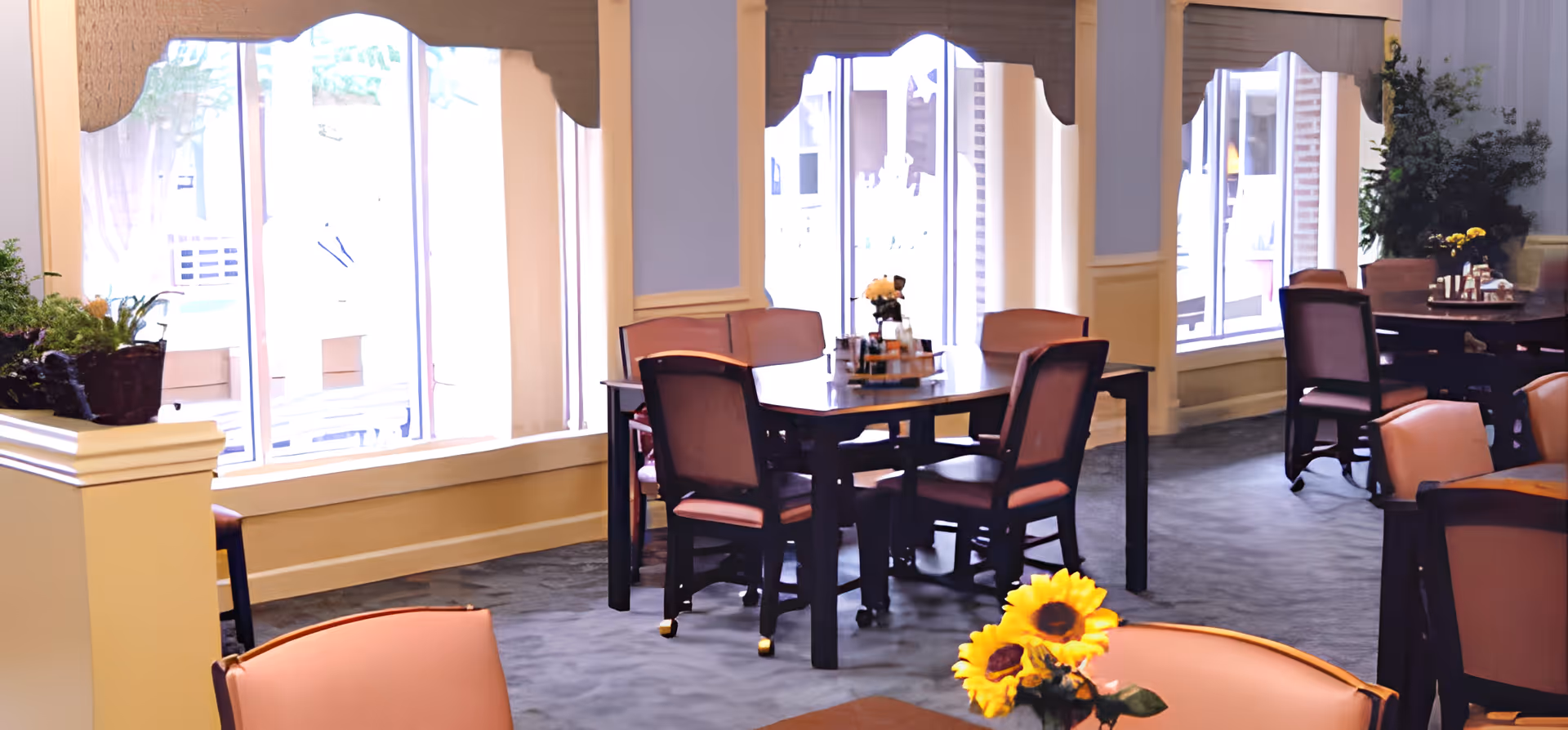 Interior view of a dining area with several tables and chairs arranged near large windows. The room is decorated with plants and flowers, including a vase with sunflowers on a table in the foreground. The space has soft lighting and a calm, welcoming atmosphere.