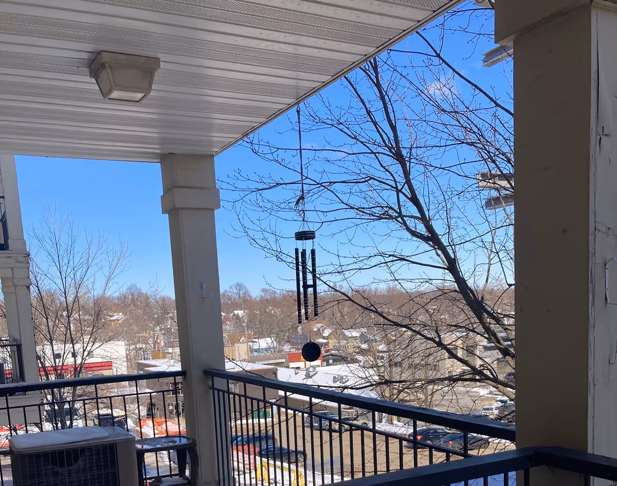 View from a covered balcony with railing and wind chimes overlooking a snowy parking lot, trees, and nearby buildings under a clear blue sky.