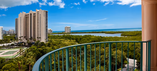 View from a curved balcony railing over treetops toward high-rise buildings and a waterfront under a blue sky.
