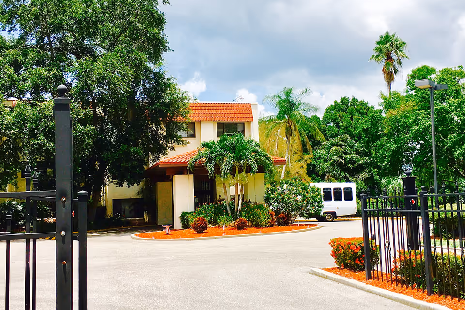 Exterior view of a building with a red-tiled roof surrounded by lush green trees and palm trees, with a driveway and a white van parked near the entrance. Black metal gates and landscaped bushes with red mulch are visible in the foreground under a partly cloudy sky.