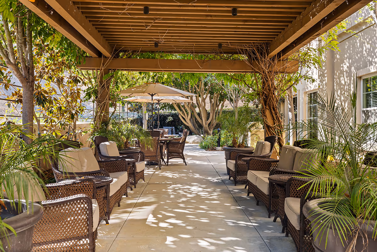 Outdoor covered patio area with cushioned wicker chairs arranged along both sides of a walkway. There are large potted plants and trees providing shade, and tables with umbrellas are visible further down the path.