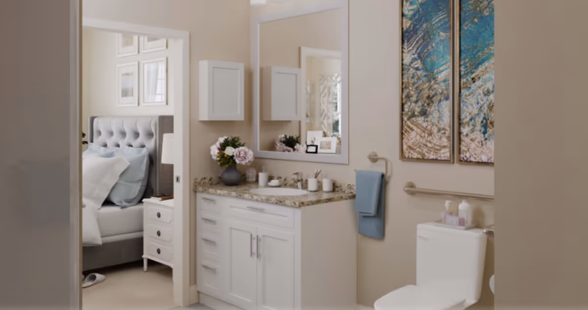 A light, accessible bathroom with a vanity and mirror, toilet with grab bar and towel, and a view into a bedroom.