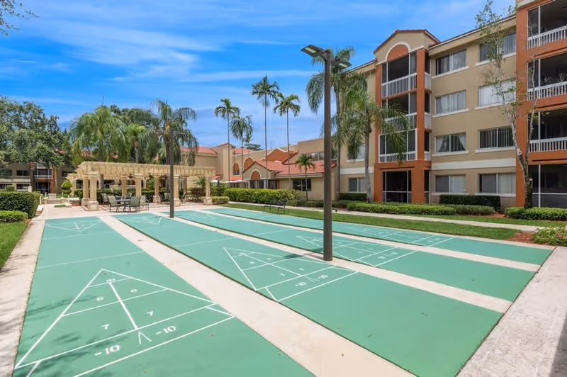 Outdoor shuffleboard courts with green playing surfaces and white markings, surrounded by palm trees and landscaping. In the background, there is a pergola with tables and chairs, and a multi-story residential building with balconies and windows under a blue sky.