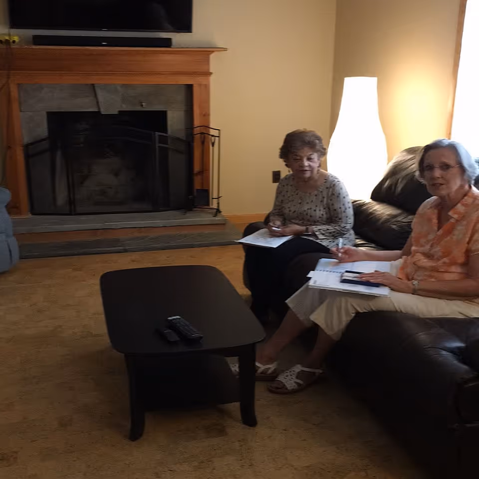 Two elderly women sitting on a dark leather couch in a living room with a fireplace and a tall white lamp. They are holding papers and pens, appearing to be engaged in a discussion or activity. A black coffee table with remote controls is in front of them.