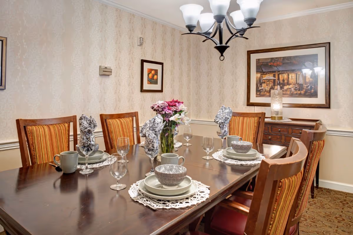 A formal dining room set for six with a dark wooden table, striped-upholstery chairs, a central floral centerpiece, chandelier, and framed artwork on patterned wallpaper.