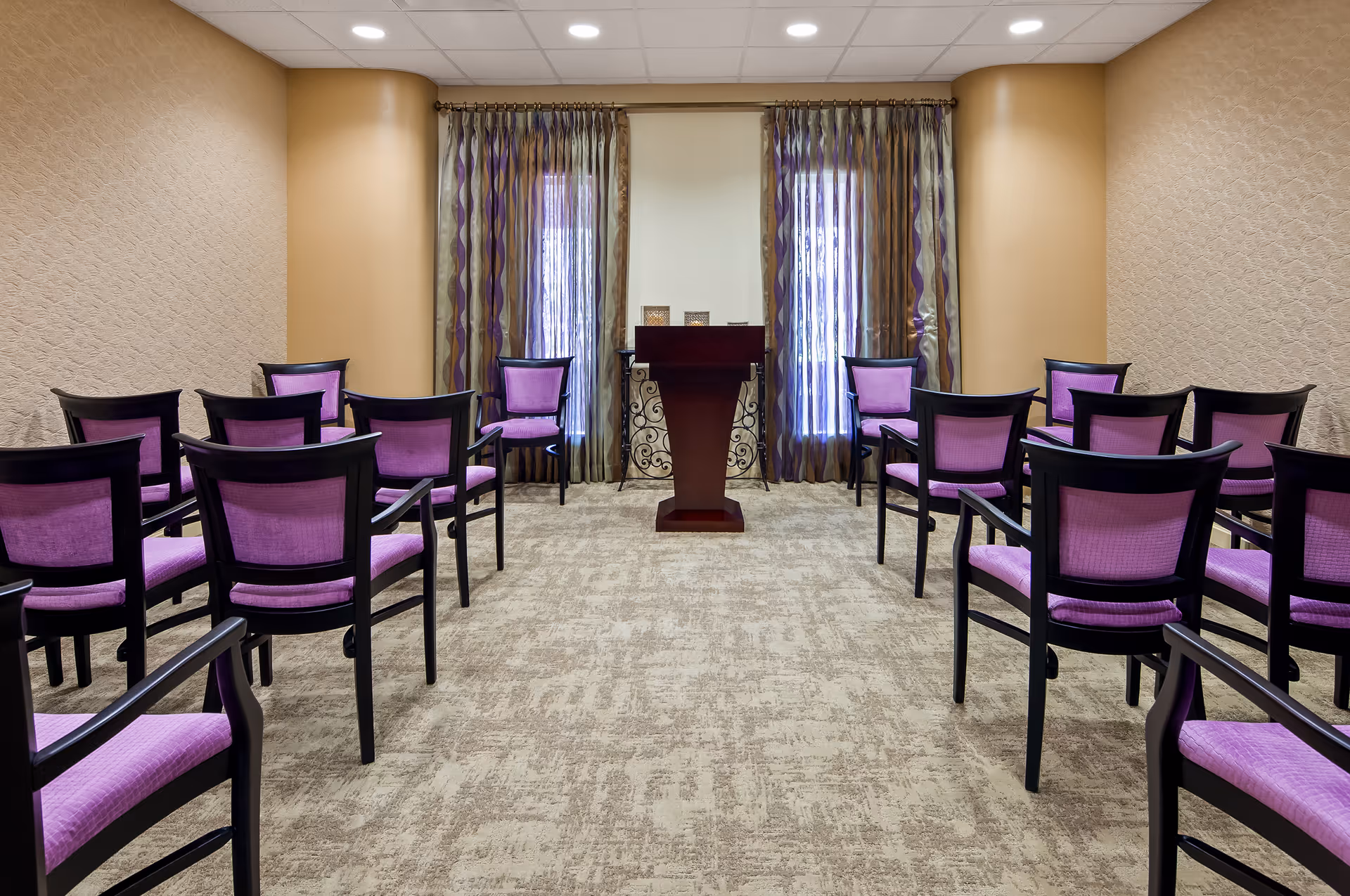 A small conference or meeting room with rows of black-framed chairs featuring purple cushions arranged facing a wooden podium. The room has beige walls with textured wallpaper, two tall windows with sheer and patterned curtains, and a beige carpeted floor.