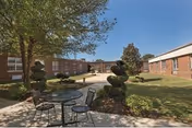 Outdoor courtyard area at a senior living facility with a round metal table and four chairs on a concrete patio. The courtyard is surrounded by single-story brick buildings with windows. There are manicured bushes, a tree providing shade, and a clear blue sky.