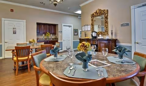 A dining area in a senior living facility with a round table set for four people, including plates, silverware, glasses, and folded napkins. The room features wooden chairs, a sideboard with a large ornate mirror above it, and a small kitchenette area in the background with cabinets and a countertop. The walls are beige and the floor is wooden.