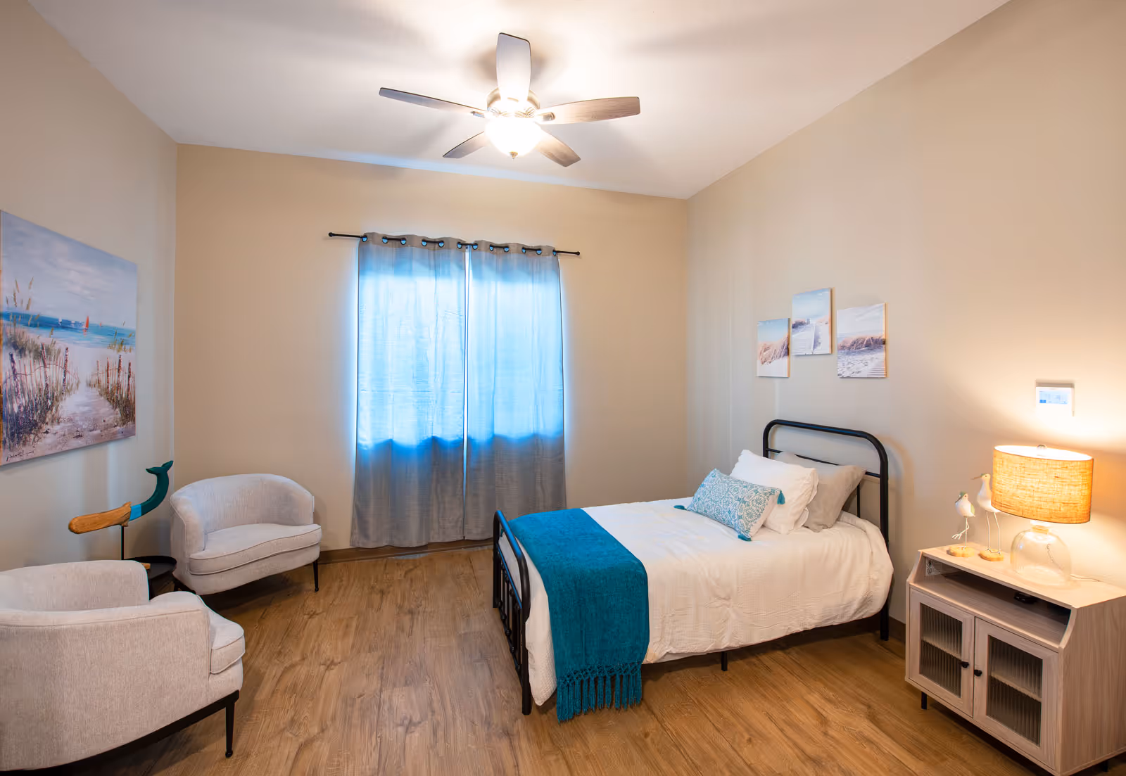 A cozy bedroom with a single bed featuring white bedding and a teal throw blanket. There are two light gray armchairs in the corner, a wooden nightstand with a lamp, and three small beach-themed pictures on the wall above the bed. A ceiling fan with a light is mounted on the ceiling, and a window with blue curtains lets in soft natural light.