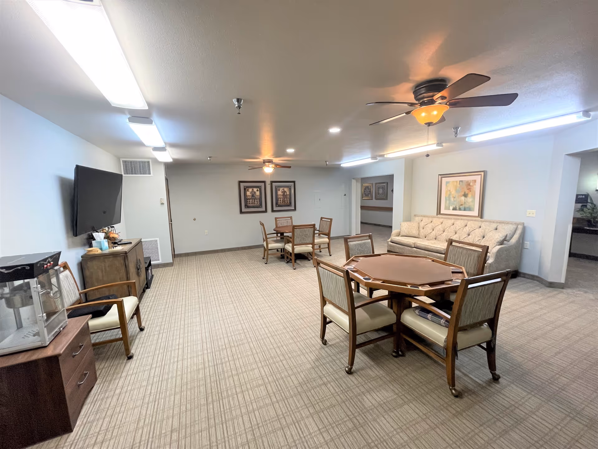A spacious common area featuring a round wooden table surrounded by chairs, a couch, and a television mounted on the wall. The room is well-lit with ceiling lights and has a neutral color scheme.