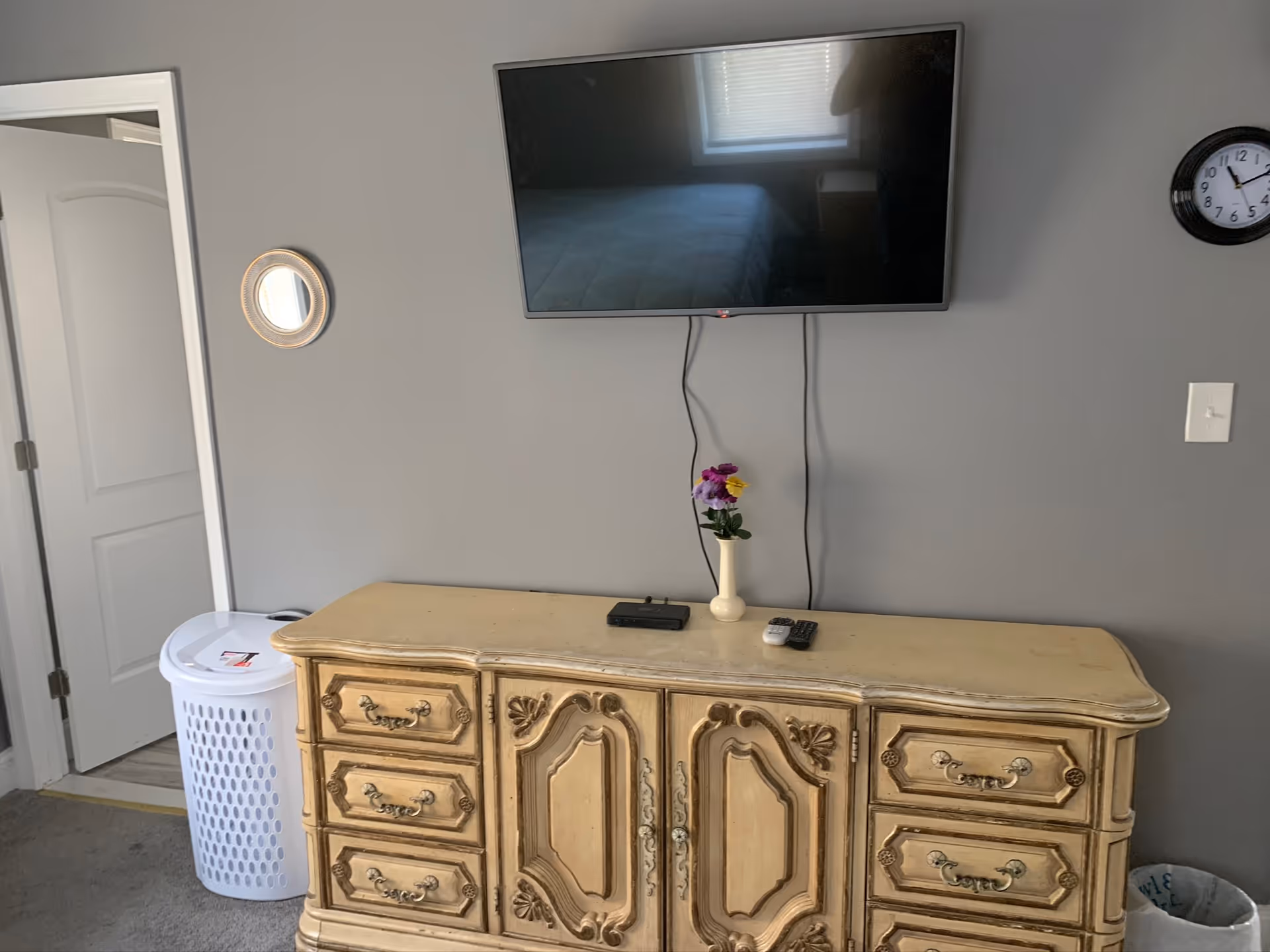 A beige ornate dresser with six drawers and two cabinet doors sits against a gray wall. Above it is a mounted flat-screen TV with visible cords hanging down. On top of the dresser are a small vase with purple and yellow flowers, a remote control, and a small black device. To the left of the dresser is a white laundry basket near an open door, and to the right is a trash can. A round clock and a light switch are mounted on the wall.