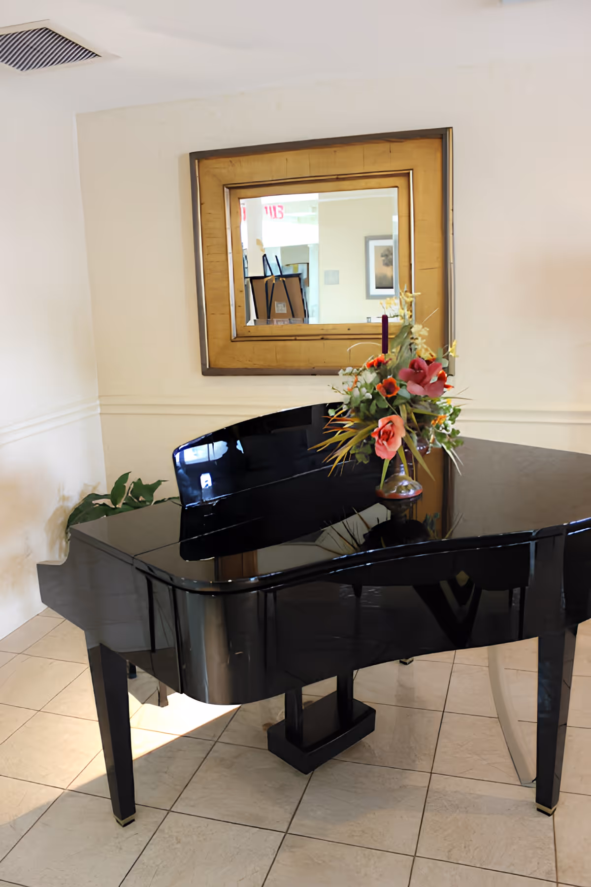 A glossy black grand piano with a floral arrangement on top, placed in a tiled room with cream-colored walls. Above the piano is a large square mirror with a gold frame reflecting part of the room behind.