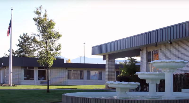 Outdoor view of a senior living facility with a white brick building, a flagpole with an American flag, a tree, and a multi-tiered white fountain in the foreground.