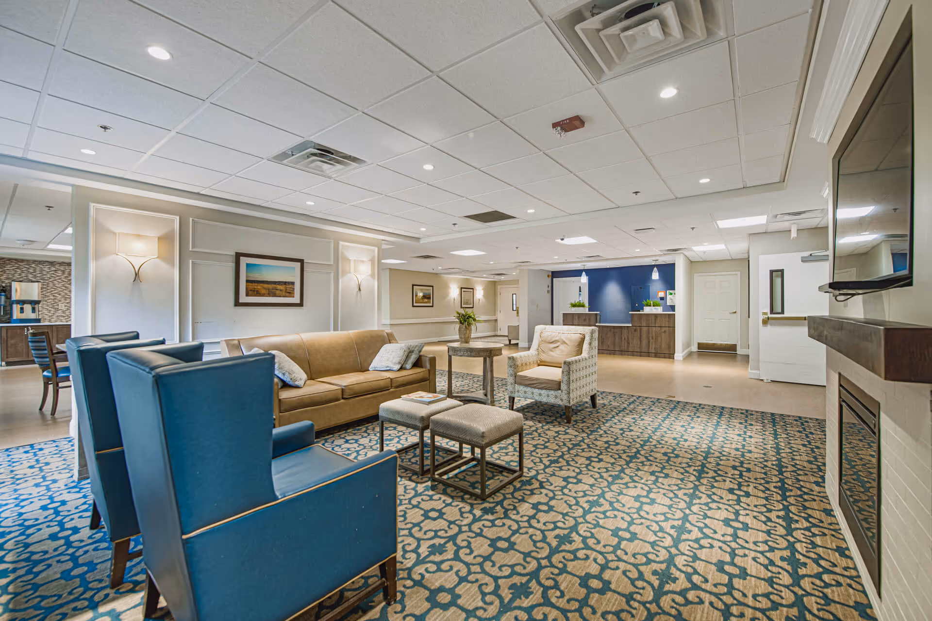 A spacious and well-lit common area in a senior living facility featuring a seating arrangement with a tan sofa, two blue armchairs, a patterned armchair, two small ottomans, and a round wooden table. The floor is covered with a blue and beige patterned carpet. The room has a white ceiling with recessed lighting and a fireplace with a mounted TV above it. In the background, there is a reception desk with a blue accent wall and pendant lights, as well as framed artwork on the walls.