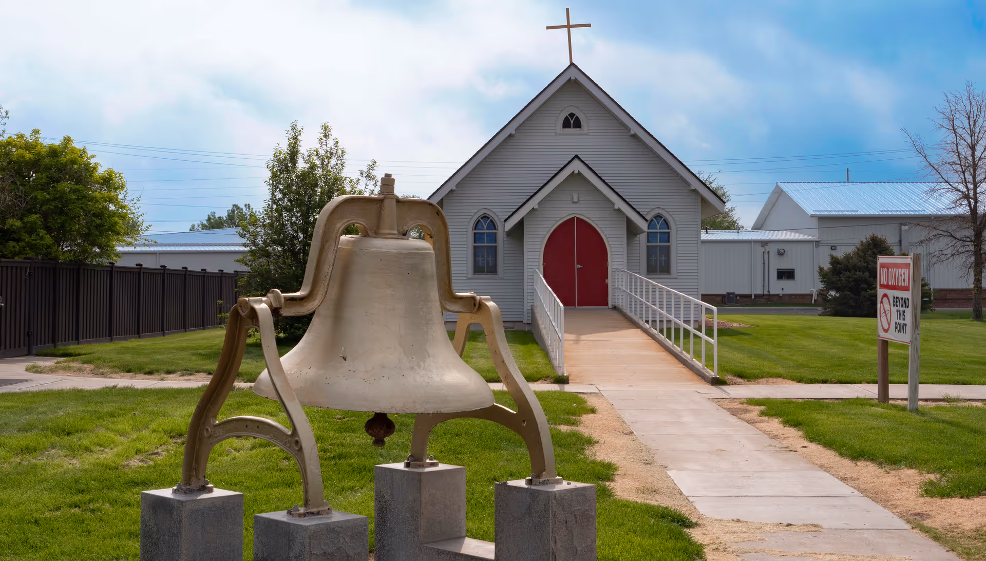 A small white chapel with red double doors and a cross on the roof, with a large bell mounted on pedestals in the grassy foreground.