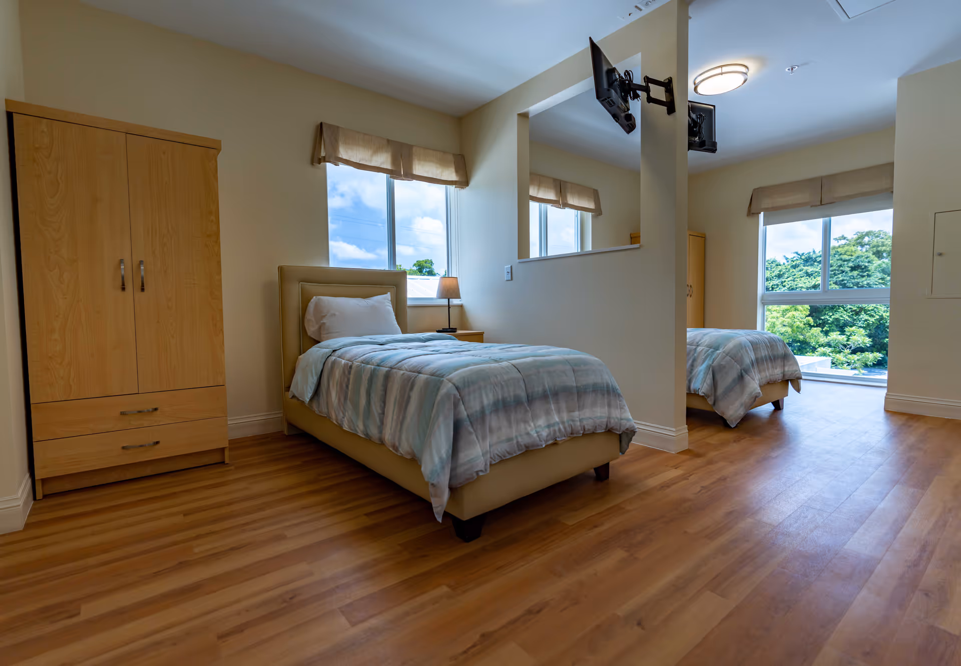A bright and clean bedroom in a senior living facility featuring two single beds with light blue and white bedding, a wooden wardrobe, a small bedside table with a lamp, large windows with beige valances letting in natural light, and two wall-mounted televisions.