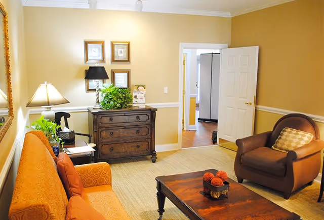 A cozy living room with an orange patterned sofa and a brown armchair with a checkered pillow. A wooden coffee table with decorative items sits in the center on a beige rug. Against the wall is a dark wooden chest of drawers with a black lamp, green plants, and framed pictures above it. The walls are painted beige with white trim, and an open door leads to another room with wooden flooring.