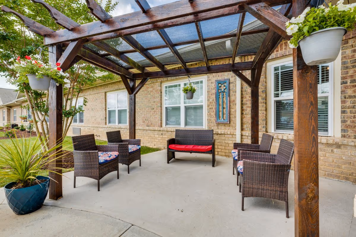 Outdoor seating area at Chatham Ridge Assisted Living featuring a wooden pergola with a translucent roof, six wicker chairs with cushions, and a wicker loveseat with a red cushion. The area is decorated with hanging flower pots and potted plants, adjacent to a brick building with windows.