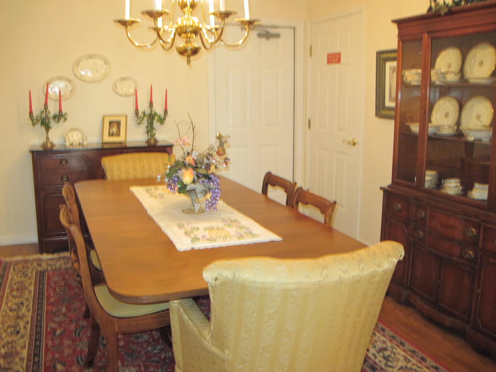 A traditional dining room with a wooden dining table surrounded by six chairs, including two upholstered armchairs at the ends. The table has a decorative runner and a floral centerpiece. On the right side, there is a wooden china cabinet displaying plates and bowls. On the left side, a sideboard holds two candelabras with red candles and a framed picture. A brass chandelier hangs above the table, and the floor is covered with a patterned rug.