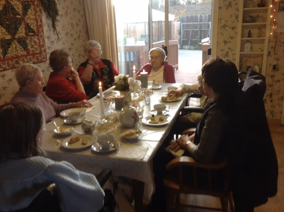 A group of elderly women sitting around a dining table set with tea cups, plates with snacks, candles, and a floral centerpiece in a cozy room with floral wallpaper and a sliding glass door leading outside.