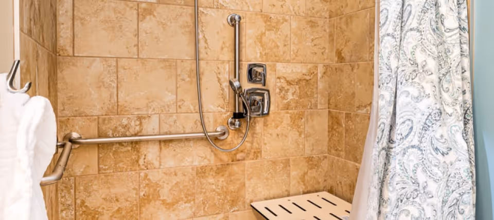 Tiled accessible shower with grab bars, a handheld showerhead, and a bench partially visible behind a patterned shower curtain.