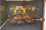 A dining room with a wooden table and six matching chairs. The walls are painted green and decorated with framed artwork, including a rooster and various rustic-themed pictures. There is a chandelier hanging above the table and two table lamps on a sideboard against the back wall.